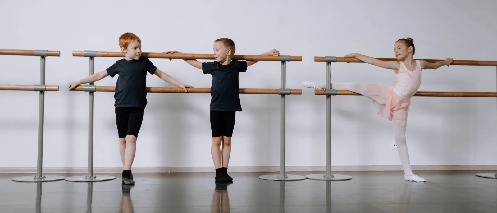 Two boys and a girl at the barre in a ballet class, the boys in black t-shirts and shorts